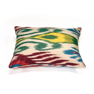 A rectangular ikat cushion showcasing a colourful pattern of red, green, blue, and yellow abstract shapes on a cream background. The design features the characteristic blurred edges of ikat weaving.