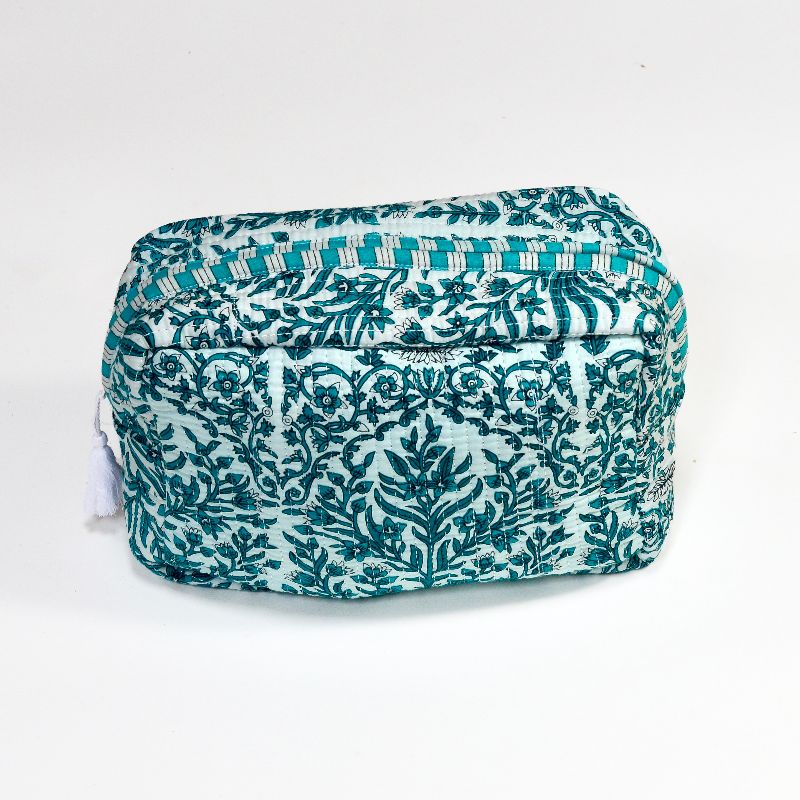A handcrafted cosmetics bag with a light teal fabric cover, featuring an intricate teal floral and vine embroidery pattern, accented by a teal and white striped border and a white tassel, set against a plain white background.