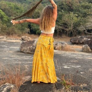 Full back view of a long-haired tattooed woman standing on rocky terrain, wearing a high-waisted marigold yellow wrap maxi skirt and holding up an olive green scarf.
