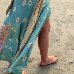 Side view of a barefoot woman walking on a sandy beach wearing a fluid turquoise silk wrap maxi skirt with diamond patterns.