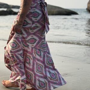 Side profile of a barefoot, tattooed woman walking along a sandy shoreline wearing a sweeping magenta and sage green ikat maxi wrap skirt.