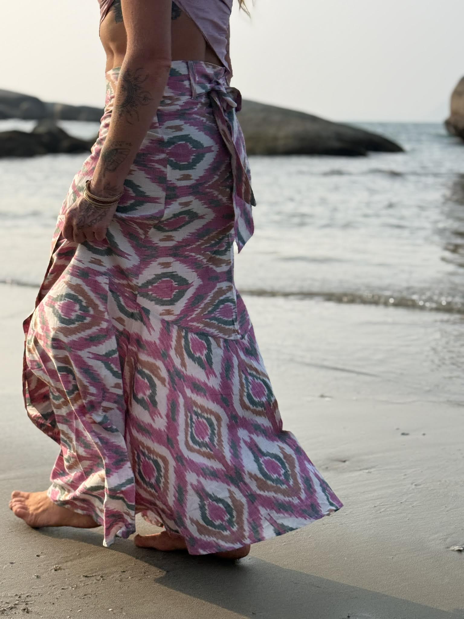 Side profile of a barefoot, tattooed woman walking along a sandy shoreline wearing a sweeping magenta and sage green ikat maxi wrap skirt.