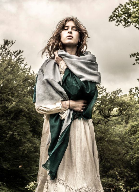 A low-angle portrait of a woman looking downward, wrapped in a two-tone grey and deep emerald green wool shawl over a cream-colored dress with lace cuffs, set against an outdoor background of green trees and a moody, overcast sky.