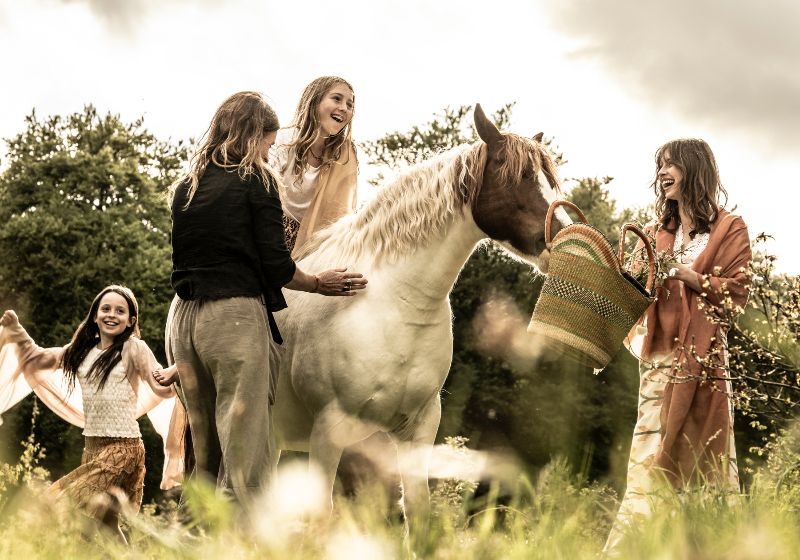 A joyful, candid outdoor scene featuring a group interacting with a white and brown pinto horse in a grassy field. One person sits on the horse while another stands nearby, as a third person laughs holding a woven market basket toward the horse, and a fourth person dances happily in the background.