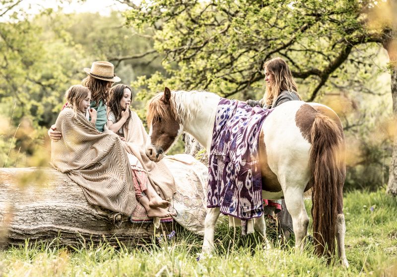 A woman wearing a straw hat sits on a large fallen log alongside two young girls wrapped in textured beige shawls, looking toward a white and brown pinto horse draped in a purple and white patterned blanket, while another woman stands nearby in a sunny pasture.