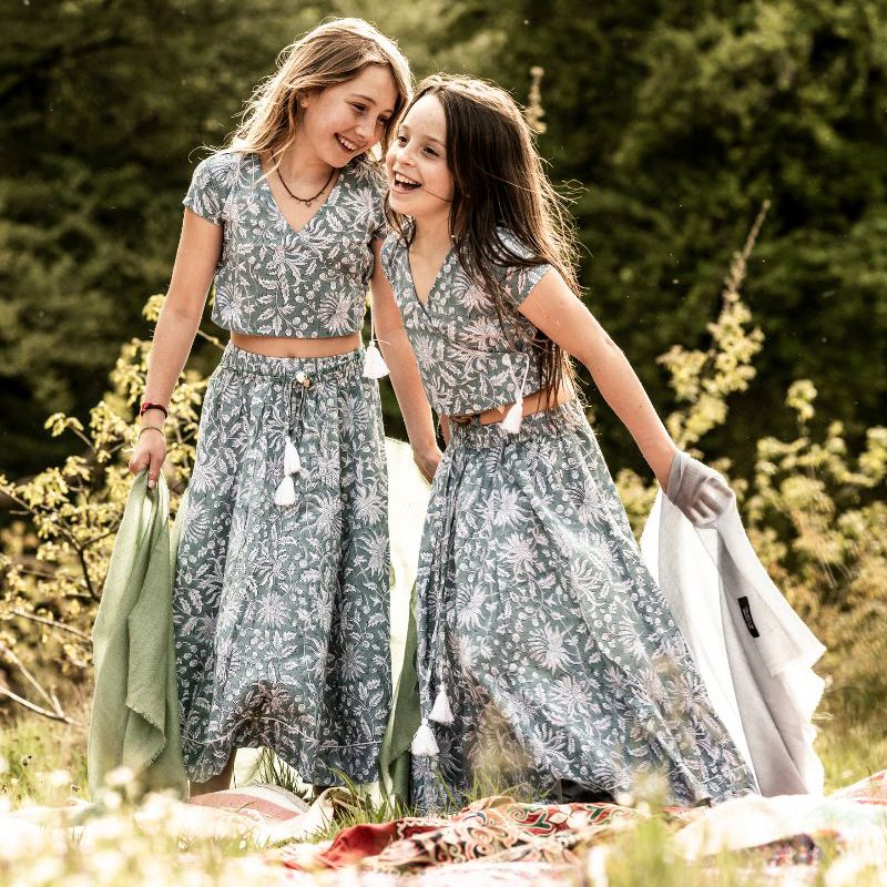 Two young girls laugh together while standing on a patterned blanket in a grassy field, wearing matching bohemian two-piece outfits consisting of a short-sleeved wrap crop top and a long maxi skirt in a blue and white floral block print, while holding lightweight sage green and grey fabric shawls.