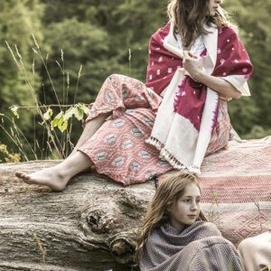A woman in a straw hat loungeing on a log, showcasing the fluid maxi drape of the rose-toned ikat wrap skirt alongside a child wrapped in a woven blanket in a grassy field.