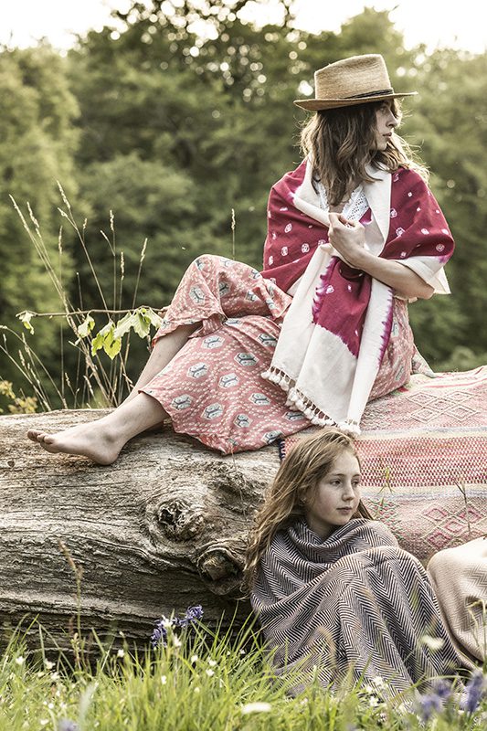A woman in a straw hat loungeing on a log, showcasing the fluid maxi drape of the rose-toned ikat wrap skirt alongside a child wrapped in a woven blanket in a grassy field.