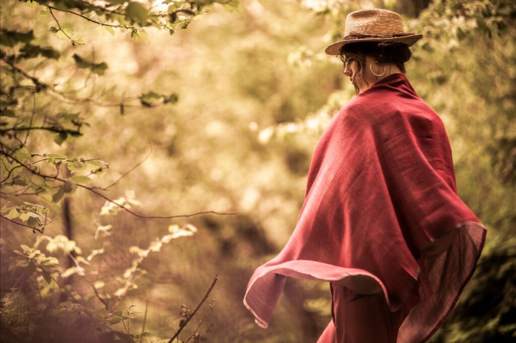 A woman in profile wearing a straw hat and large hoop earring, looking down while draped in a flowing, textured dark pink or raspberry-colored shawl, set against a softly blurred, warm-toned forest or woodland background.
