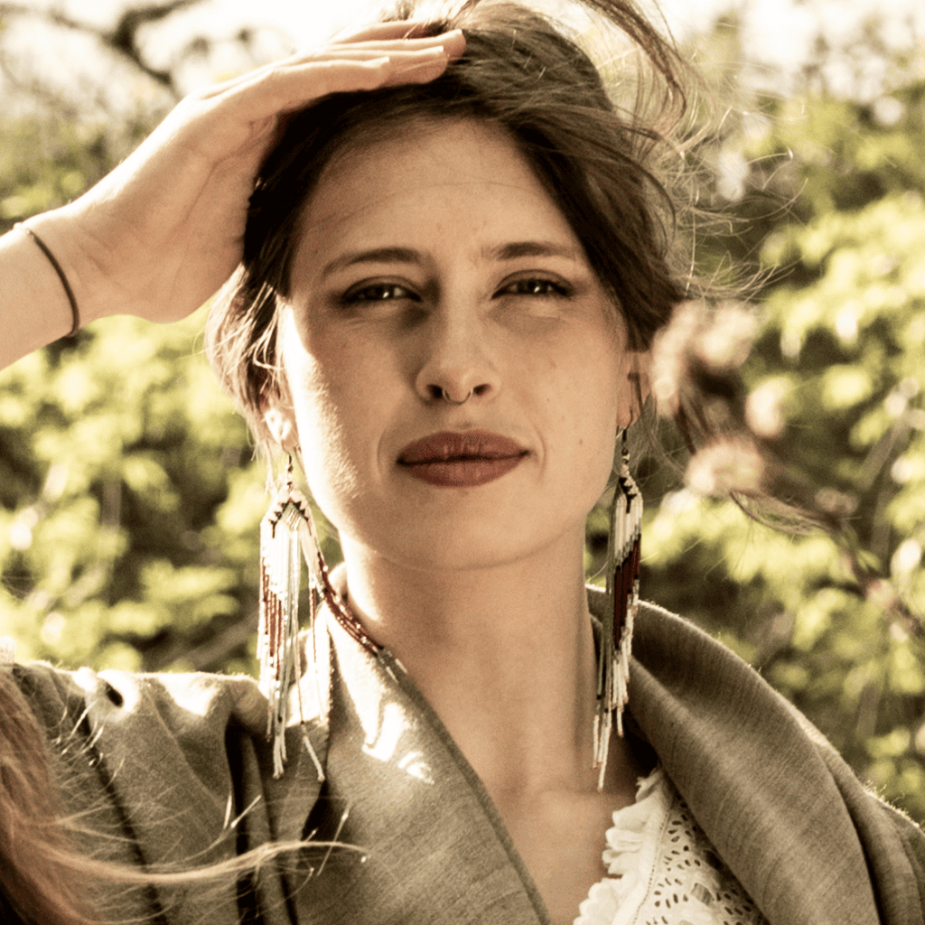 Close-up portrait of a woman outdoors wearing long, bohemian-style beaded fringe earrings in shades of turquoise, burgundy, and white. She has a hand raised to her forehead against a soft-focus background of green foliage in warm, natural sunlight.