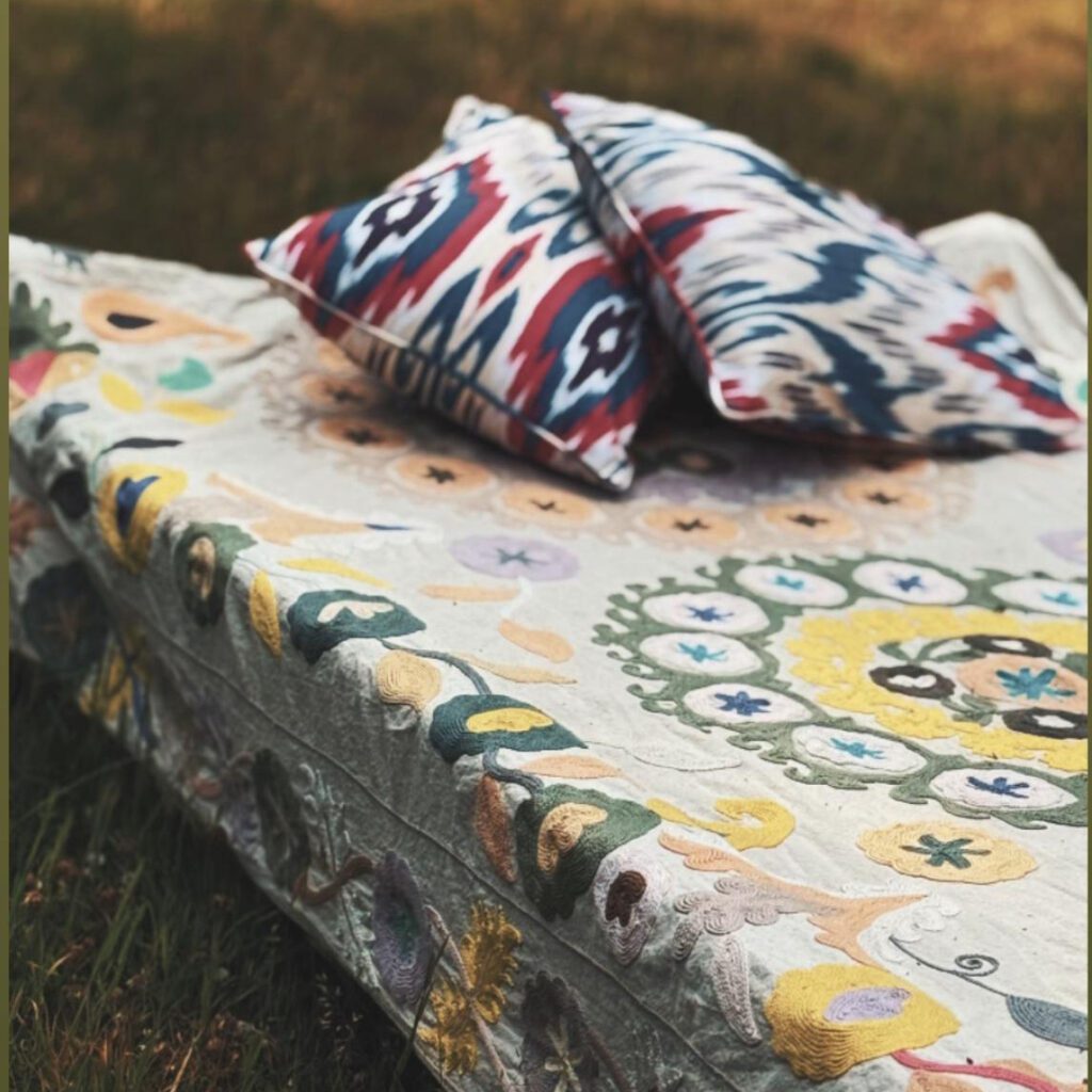 An outdoor setup on green grass featuring an intricately embroidered cream suzani quilt or bedspread with vibrant yellow, green, and red circular floral patterns, topped with two decorative silk ikat cushions in striking red, blue, and white geometric designs.