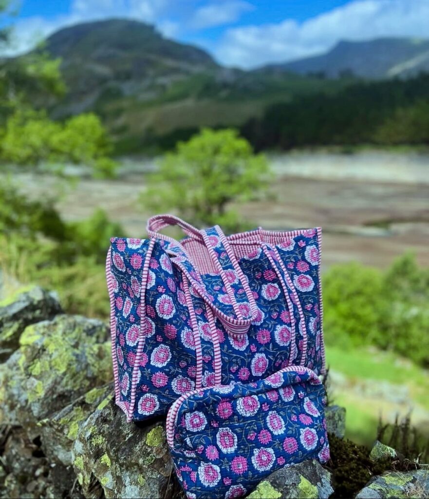 A matching tote bag and small zippered pouch crafted from blue organic fabric with pink and white floral block print patterns. The bags feature pink and white striped piping and handles, resting outdoors on a mossy stone wall with a scenic, soft-focus mountain landscape and green hills in the background.