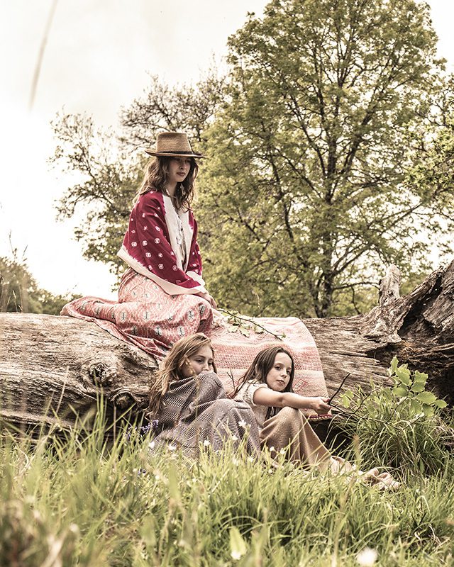 A woman in a straw hat sits on a large fallen log in a grassy field wearing a pink ikat wrap skirt and berry red shawl, while two young girls sit in the grass below her wrapped in woven blankets.