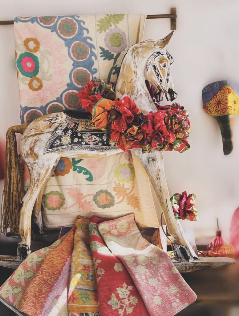 A decorative vintage wooden rocking horse adorned with vibrant, ruffled fabric garlands stands in front of a hanging suzani textile with colorful floral embroidery. In the foreground, several folded kantha quilts and silk fabrics in warm shades of pink, red, and orange are neatly draped, showcasing detailed patterns.