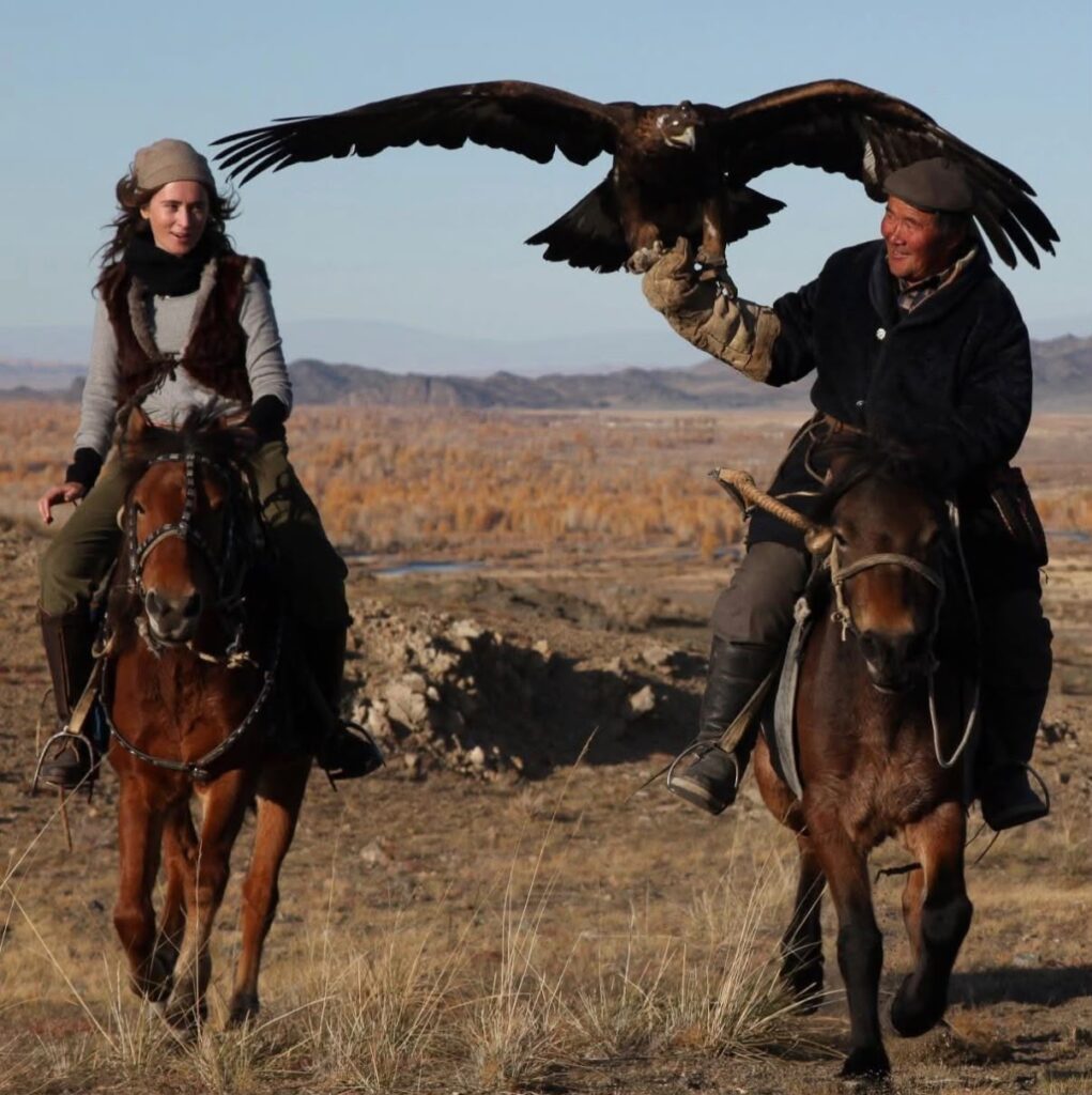 Beshlie McKelvie on horseback alongside a Kazakh eagle hunter holding a golden eagle with wings spread across the Mongolian steppe.